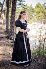 Fototapeta premium Young, beautiful girl in a black medieval dress walks through the gloomy pine forest.
