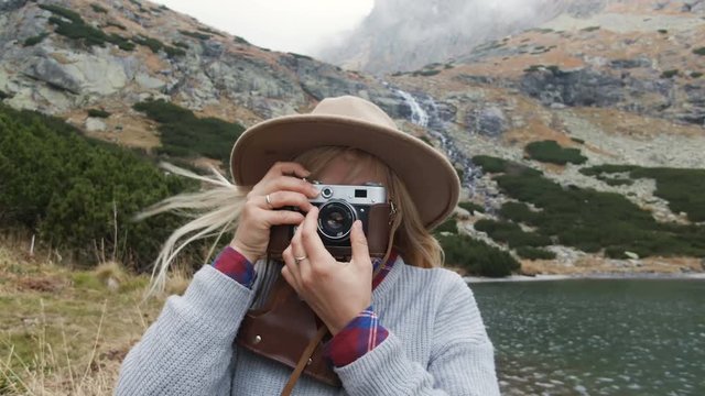 Girl traveler in a hat and a vintage film camera in her hands. Photographs mountain landscapes on an old film camera. On the shore of  mountain lake.