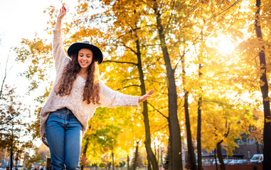 Joyful stylish young woman with raised arms walking on colorful autumn city street at sunny day