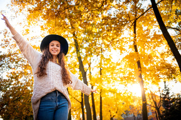 Joyful stylish young woman with raised arms walking on colorful autumn city street at sunny day