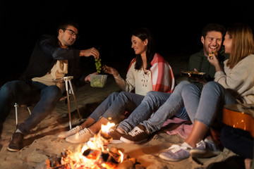 leisure and people concept - group of smiling friends having picnic at camp fire on beach at night