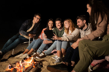 leisure and people concept - group of smiling friends sitting at camp fire with tablet pc computer on beach at night