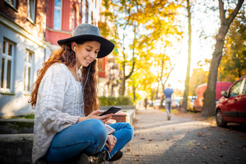 Beautiful stylish woman sitting on street with crossed legs at autumn day and using mobile phone