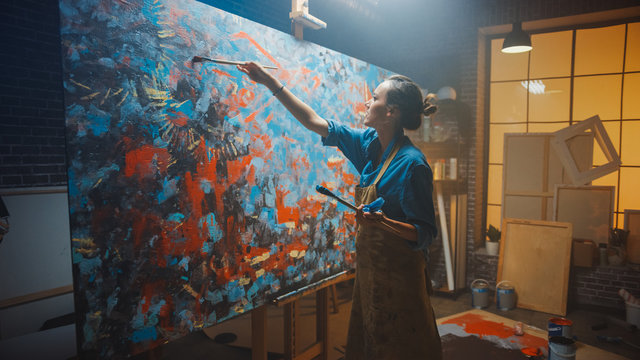 Talented Female Artist Works on Abstract Oil Painting, Using Paint Brush She Creates Modern Masterpiece. Dark and Messy Creative Studio where Large Canvas Stands on Easel Illuminated