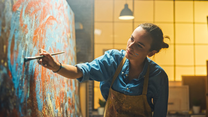 Portrait of Talented Female Artist Working on a Modern Abstract Oil Painting, Gesturing with Broad Strokes Using Paint Brush. Dark Creative Studio Large Picture Stands on Easel Illuminated. - obrazy, fototapety, plakaty