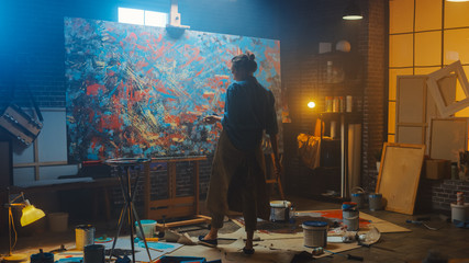 Talented Female Artist Works on Abstract Oil Painting, with Broad Strokes of Paint Brush She Creates Modern Masterpiece. Dark and Messy Creative Studio where Large Canvas Stands on Easel Illuminated 
