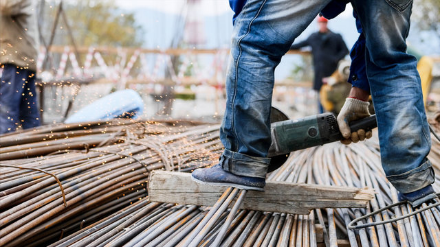 Close-up view of legs in foreground of a worker man in blue jeans, cutting rebar with a saw blade at construction site. Right leg is on the metal tubes which are on wooden stud, sparks from process.