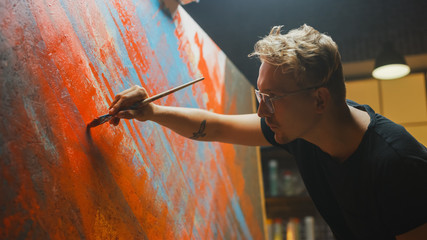 Portrait of Artist Working on Abstract Painting, Uses Paint Brush To Create Daringly Emotional Modern Picture. Dark Creative Studio Large Canvas Stands on Easel Illuminated. Side View Close-up Shot