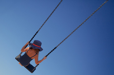 Girl in a swimsuit and hat swinging on a swing against a blue sky