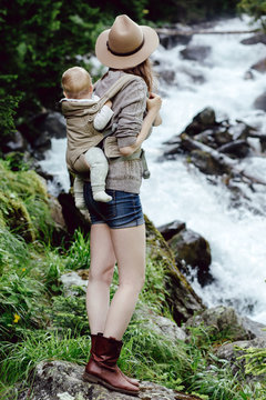 Preselect Julia! Mother with baby in carrier stand near waterfall in coniferous forest
