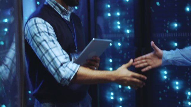 Two IT engineers walking in working rack server corridor inspecting modern computer data center. Close-up business partners shaking hands of successful deal.