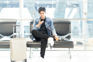 Portrait of a young Asian man waiting for departure at the airport while using his phone and drink hot coffee.