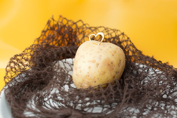 We love potatoes concept. Closeup of potato with small enamel heart on brown net bag in front of yellow background