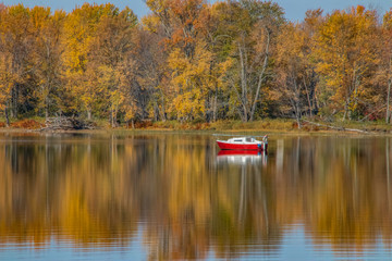 Small red sailboat moored off shore, calm water with autumn leaves and trees, nobody