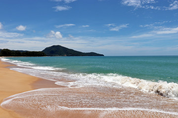 Tropical sandy beach with a frothy wave, scenic view to empty sea coast with yellow sand, azure water and mountains. Picturesque seascape with blue sky and white clouds