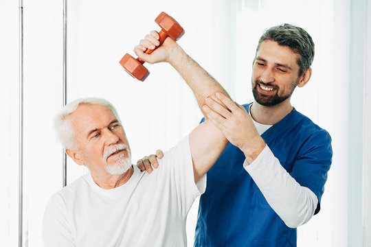 Positive physiotherapist helping his senior patient to lift a dumbbell. Physio treatment at rehab center