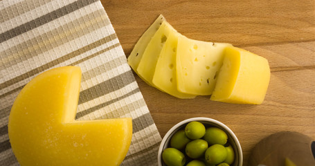 Cheese wheel with a cut piece on a wooden Board. Green olives and pieces of cheese nearby. The view from the top