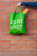 Green eco bag with a white text zero waste in the hand on a background of a brick wall with copy...