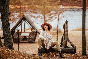 Portrait of beautiful brunette woman sitting near the lake, wooden arbor and stairway. Fashion outdoor shot of pleased lady in cozy clothes and straw hat enjoying nature views in autumn countryside.