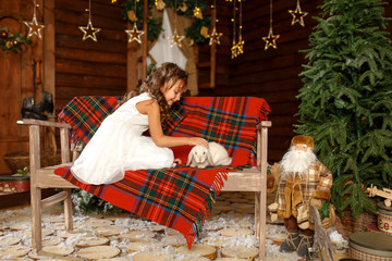 New Year 2020. Merry Christmas, happy holidays. A little girl in white dress sitting on the bench with white rabbit. Magic Light in Night Xmas Tree Interior