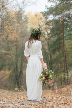 bride in a white dress with a bouquet and a wreath stands against the background of an autumn forest