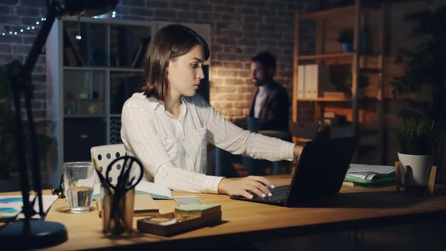 Exhausted girl is working with laptop when manager is bringing more papers at night in dark office, woman is feeling unhappy and stressed. People and job concept.