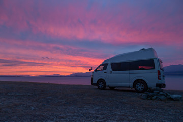 A campervan is standing at Lake Tekapo, New Zealand, looking into the sunset. The sky is scattered with some clouds with vibrant red, purple and orange colors.
