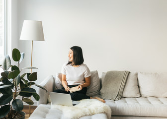 Smiling Asian female sitting on sofa working from home in minimalist space