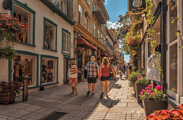 A senior couple in summer casual shorts and tops walk down rue de Petit Champlain in Quebec City's historicLower Town summer afternoon with a blue sky and clouds.