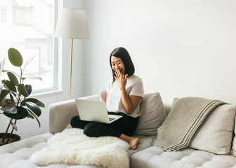Smiling Asian female sitting on sofa working from home in minimalist space