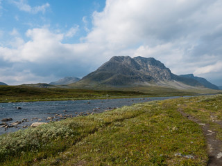 Beautiful wild Lapland nature landscape with blue glacial river, birch tree bushes, snow capped mountains Northern Sweden summer at Kungsleden hiking trail. Summer day, blue sky.