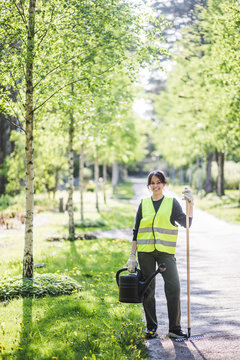 Full length portrait of smiling young woman holding watering can and rake on footpath at garden