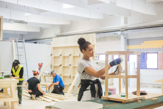 Confident young female carpenter rubbing sand paper on wooden table at workbench in illuminated workshop
