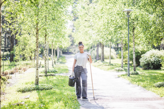 Portrait of confident young woman holding watering can and rake on footpath at garden