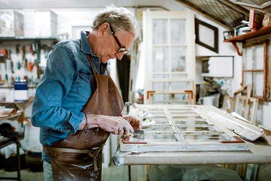 Side view of senior male owner scraping window frame on workbench at store workshop