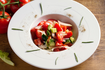 Vegetable salad of fresh tomatoes, cucumbers, bell pepper, onions and herbs in white bowl, wooden table. Horizontal image