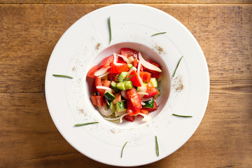 Vegetable salad of fresh tomatoes, cucumbers, bell pepper, onions and herbs in white bowl, wooden table. Overhead, horizontal image