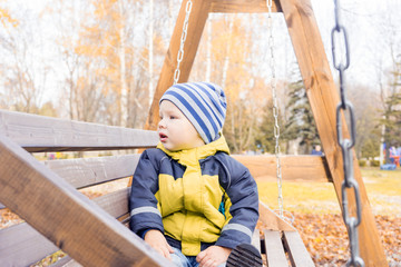 toddler child looks around with interest swinging on wooden swing on Sunny autumn day in city Park