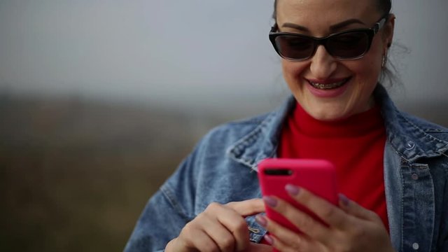 Portrait of cheerful woman with dental braces, using pink smartphone, wearing sunglasses, red sweater and blue jeans jacket.
