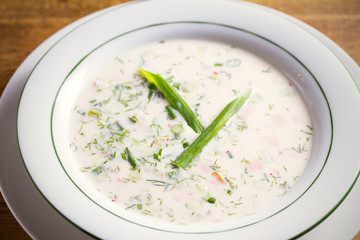 Classic cold soup Okroshka made of vegetables, boiled potatoes, eggs, and meat (beef, sausages or ham) in white bowl, wooden table. horizontal image