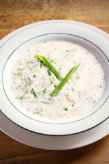 Classic cold soup Okroshka made of vegetables, boiled potatoes, eggs, and meat (beef, sausages or ham) in white bowl, wooden table. vertical image