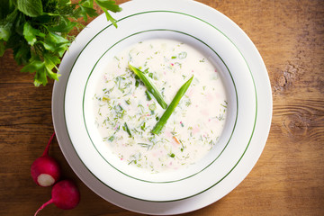 Classic cold soup Okroshka made of vegetables, boiled potatoes, eggs, and meat (beef, sausages or ham) in white bowl, wooden table. Overhead, horizontal image