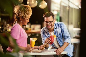 Happy couple drinking cocktails while using smart phone in a bar.