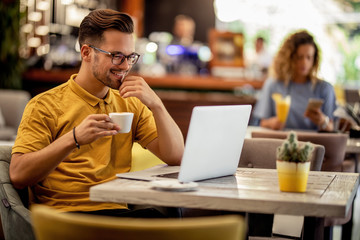 Smiling man reading an e-mail on laptop and drinking coffee in a bar.