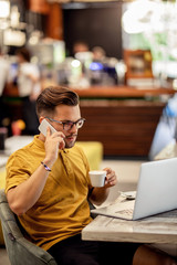 Young freelancer talking on the phone while using laptop in a cafe.