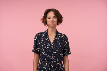 Portrait of funny young woman with short brown hair puffing out cheeks and winking at camera, being in high spirit, standing over pink background with hands down