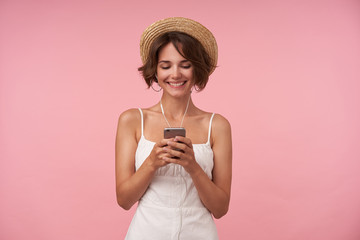 Cheerful lovely brunette female with casual hairstyle looking at screen of her phone and smiling broadly, typing message while listening to music with headphones, isolated over pink background