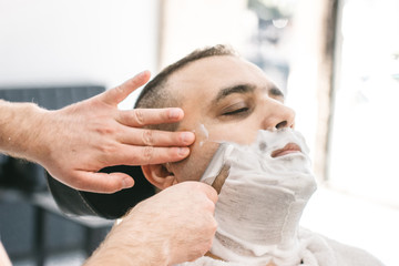 Hairdresser shaves a beard to a man with a razor in the barbershop