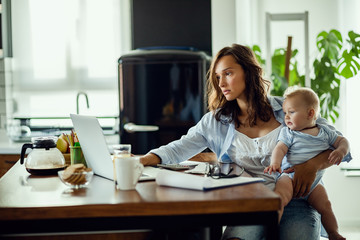 Young mother working on laptop while being with baby at home.