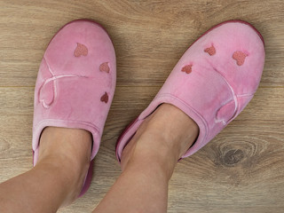 Women feet are shod in a home pink slippers. Cozy, warm and comfortable domestic shoes. On a brown wooden floor.